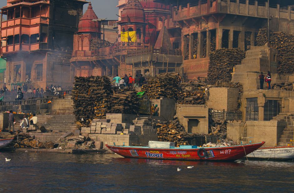 Colorful boats docked by ancient ghats with bustling urban scenery in the background.